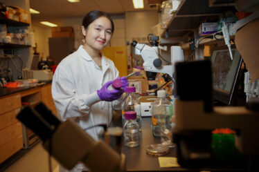Catherine Ji, in the lab with purple gloves and surrounded by microscopes and lab equipment, labels a glass bottle.