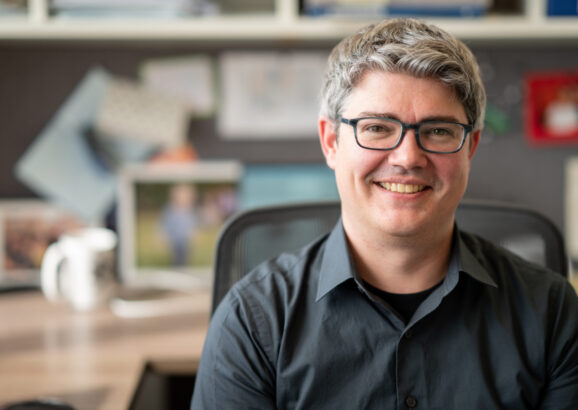 Professor Michael McDonald smiles while sitting at his desk, with blurry office background.