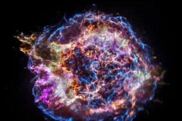 In the center of a black background is a large circular object outlined by electric blue filaments that jolt and weave throughout the structure. The object, which is the supernova remnant Cassiopeia A, includes strings of vibrant colors including purple, orange, yellow and blue.