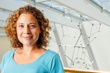 Photo of Danna Freedman indoors with a molecular sculpture hanging from the ceiling behind her
