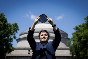 Man stands in front of the MIT dome, holding a circular wafer displaying moire patterns over his head