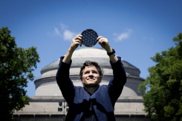 Man stands in front of the MIT dome, holding a circular wafer displaying moire patterns over his head