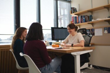 Anna-Christina Eilers sits at her desk, smiling warmly at two female students who sit facing her