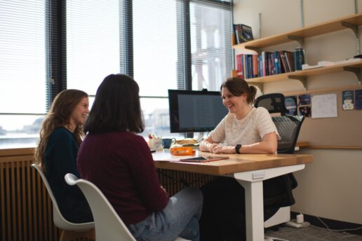 Anna-Christina Eilers sits at her desk, smiling warmly at two female students who sit facing her